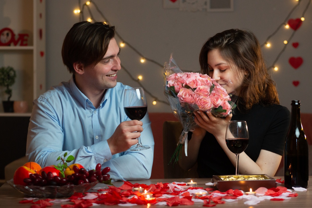 smiling-handsome-man-holding-glass-wine-looking-pretty-woman-sniffing-bouquet-flowers-sitting-table-living-room-valentine-s-day